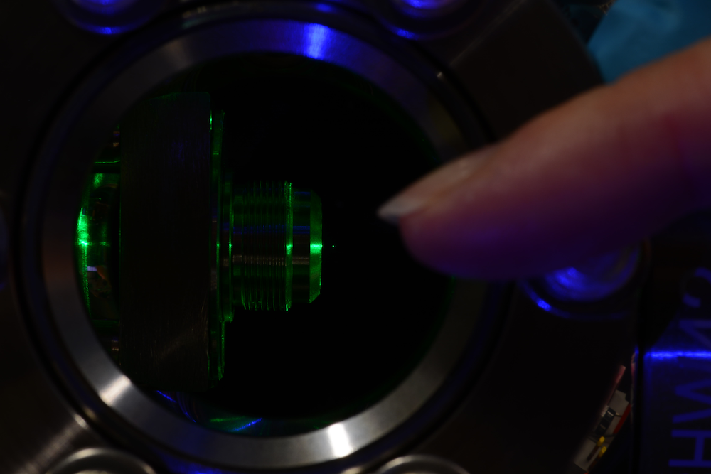 Photograph of a levitated particle. The particle is made visible by green laser light illumination. A finger, pointing at the particle, illustrates the size of the experiment.